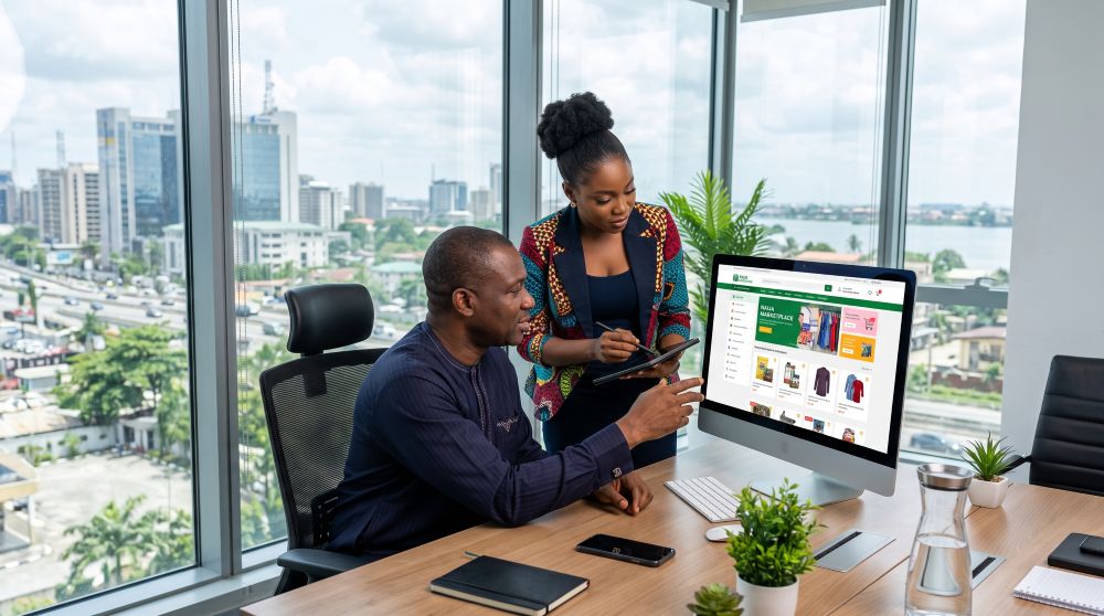 two colleagues review an online store on a large desktop monitor in a bright office with a city view outside the window, plants on the desk.